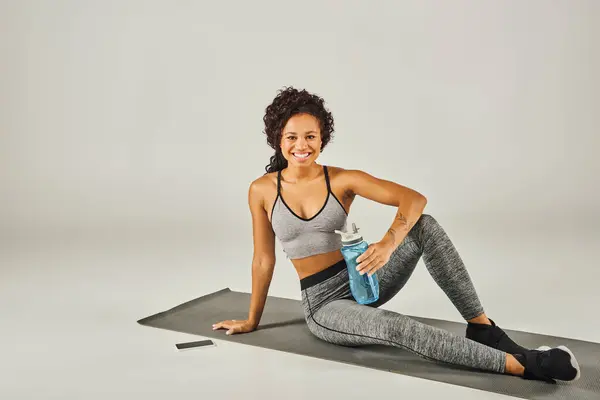 Deportista afroamericana rizada en ropa activa sentada en una esterilla de yoga, sosteniendo una botella de agua, con fondo de estudio gris. — Stock Photo