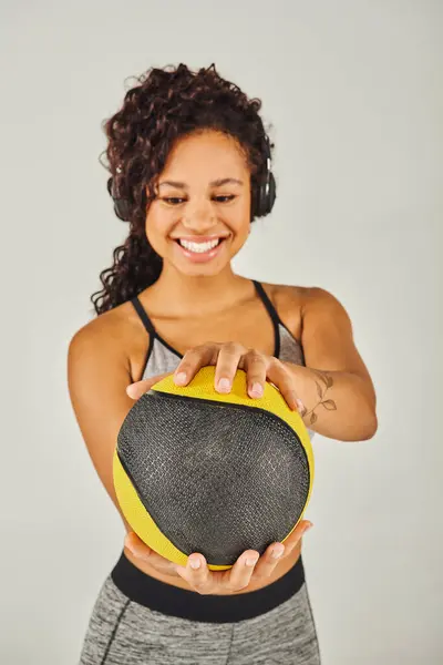 Кудрявая афроамериканская спортсменка в активной одежде энергично держит желтый и черный мячик для упражнений в студии. — стоковое фото