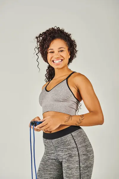 Uma esportista afro-americana encaracolada sorri enquanto segura uma corda pulando em um estúdio contra um fundo cinza. — Fotografia de Stock