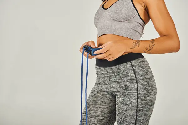 African American sportswoman in activewear energetically holds a jump rope in a studio with a grey background. — Stock Photo