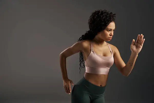 Curly African American sportswoman in sports bra and leggings striking a pose in a photo shoot with a grey backdrop. — Stock Photo