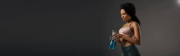 Lockige afroamerikanische Sportlerin mit einer Flasche Wasser während ihres Trainings in einem Studio mit grauem Hintergrund. — Stockfoto