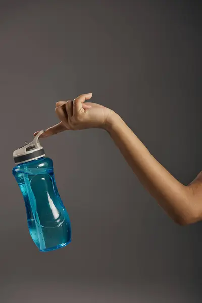 Amerikanische Sportlerin hält eine blaue Wasserflasche in einem Studio vor grauem Hintergrund. — Stockfoto