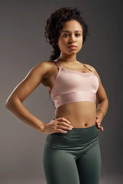 Кучерява афроамериканська спортсменка в рожевому спортивному бюстгальтері та зелених легінсах, що працюють в студії з сірим фоном. — стокове фото