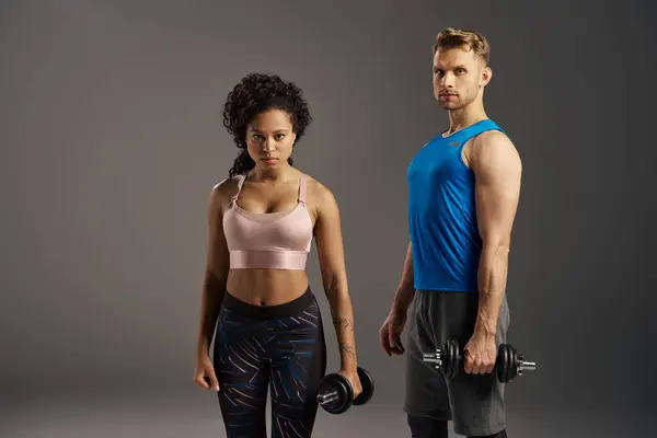 Multicultural fit couple in activewear strike poses with dumbbells against grey studio background. — Stock Photo