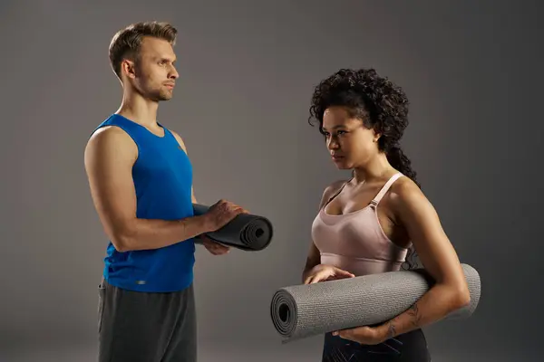 Casal multicultural em stand activewear ao lado de um tapete de ioga, pronto para uma sessão de treino em um estúdio em um fundo cinza. — Fotografia de Stock