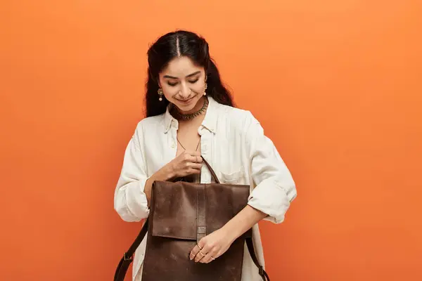Active young indian woman embraces adventure, holding a brown backpack against vibrant orange background. — Stock Photo