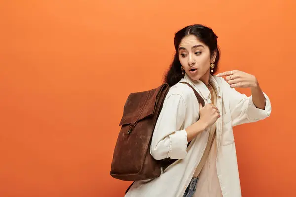 Una joven india con una mochila marrón posa sobre un fondo naranja vivo. - foto de stock