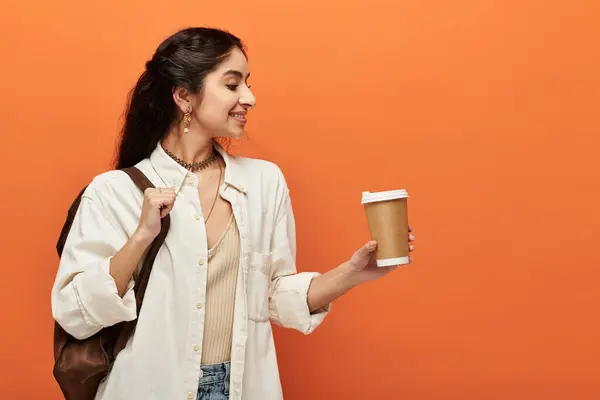 Stylish young indian woman calmly holds a cup of coffee against a vibrant orange backdrop. — Stock Photo