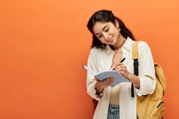 Young indian woman actively writing in notebook against bright orange background. — Stock Photo