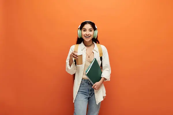 Young indian woman wearing headphones holds book against orange backdrop. — Stock Photo