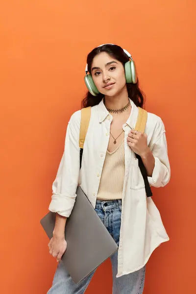 Jeune femme indienne tient énergiquement ordinateur portable tout en portant des écouteurs sur le mur orange. — Photo de stock