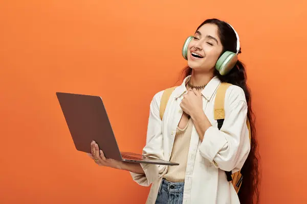 Youthful indian woman wearing headphones, typing on laptop against vibrant orange background. — Stock Photo