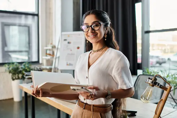 A professional indian woman with glasses holding a folder in an office setting. — Stock Photo