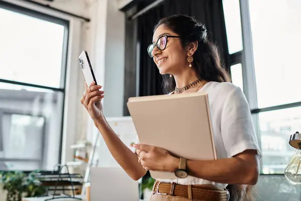 Mujer india joven en gafas sosteniendo el teléfono celular, de pie por la ventana. - foto de stock