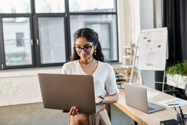 Une jeune femme indienne en lunettes utilise activement un ordinateur portable dans un cadre de bureau. — Photo de stock