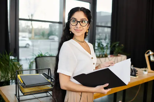 Young indian woman in chic glasses holds folder in stylish office setting. — Stock Photo
