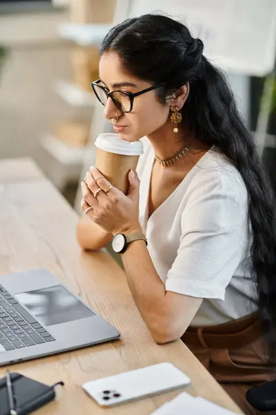 Une femme indienne professionnelle assise à un bureau avec un ordinateur portable et une tasse de café. — Photo de stock