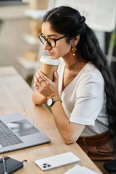 Una mujer india profesional sentada en un escritorio con una computadora portátil y una taza de café. — Stock Photo