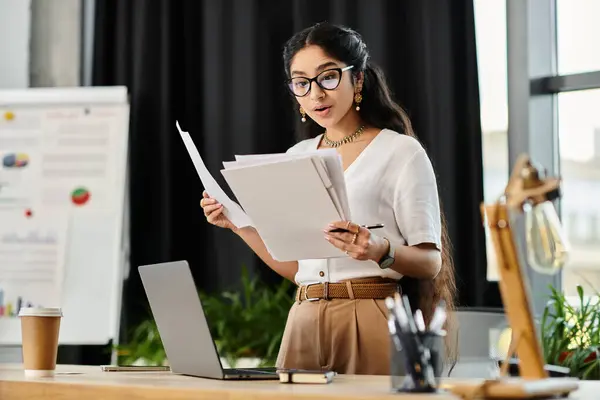 Uma mulher indiana dinâmica está diante de um laptop e papéis, envolvidos no trabalho. — Fotografia de Stock