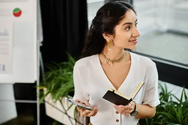 Eine indische Frau hält ein Buch in der Hand, während sie ihr Handy kontrolliert. — Stockfoto