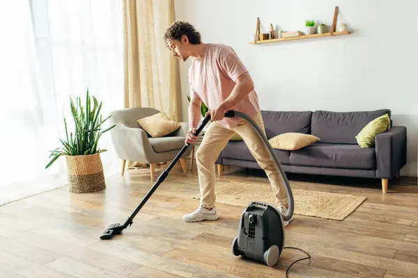 Un hombre guapo en ropa de casa acogedora meticulosamente aspira una sala de estar. - foto de stock