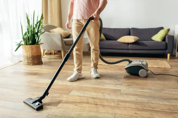 Un bell'uomo in accogliente biancheria da casa utilizza un aspirapolvere per pulire un pavimento in legno. — Foto stock