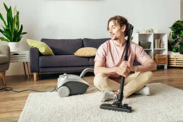 A handsome man in cozy homewear sitting on the floor while using a vacuum cleaner. — Stock Photo