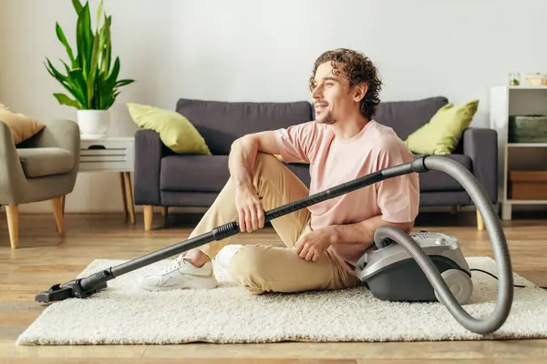 Um homem bonito sentado no chão serenamente ao usar um aspirador de pó em roupa de casa aconchegante. — Fotografia de Stock