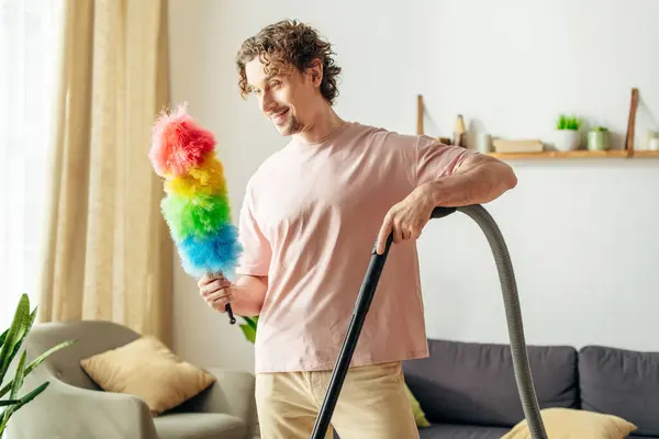 Um homem bonito em casa aconchegante alegremente segura um espanador brilhante e colorido. — Fotografia de Stock