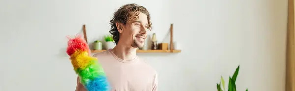 A handsome man in cozy homewear holds a multicolored duster. — Stock Photo