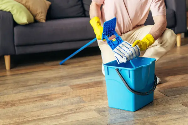 Handsome man in cozy clothes meticulously mops the living room floor. — Stock Photo