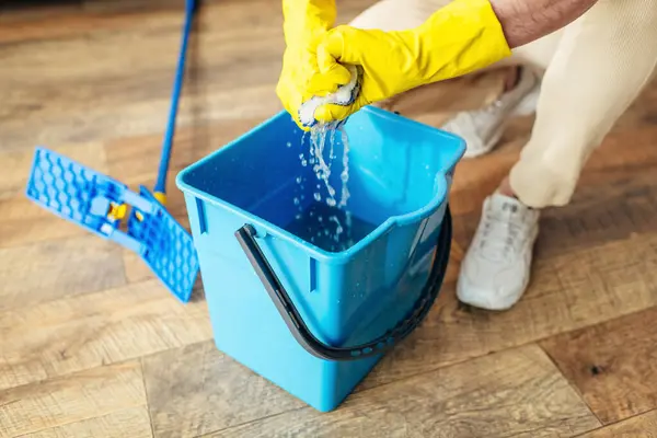 Un uomo in guanti gialli lava un secchio blu, circondato da accoglienti casalinghe. — Foto stock