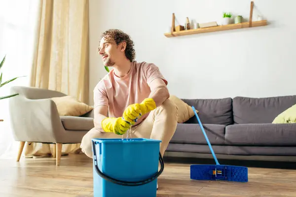 Handsome man in cozy homewear cleaning living room with mop and bucket. — Stock Photo