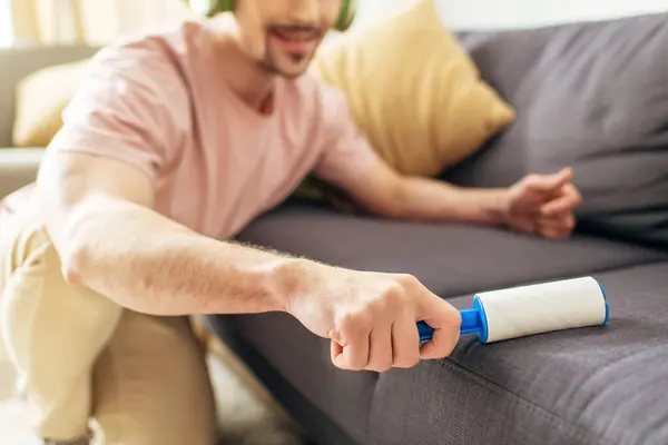 A man in cozy homewear cleaning a couch, sticky roller. — Stock Photo