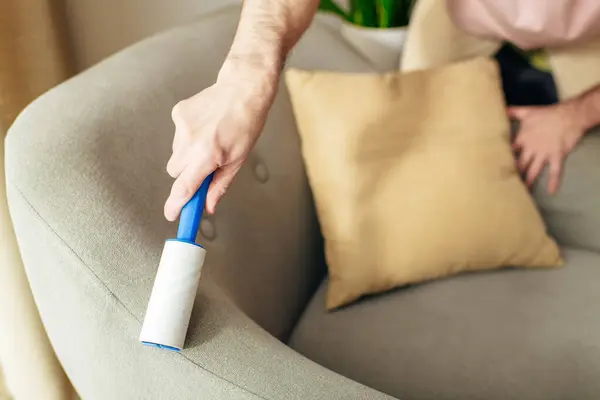 A man in cozy homewear meticulously cleans a couch using a blue sticky roller. — Stock Photo