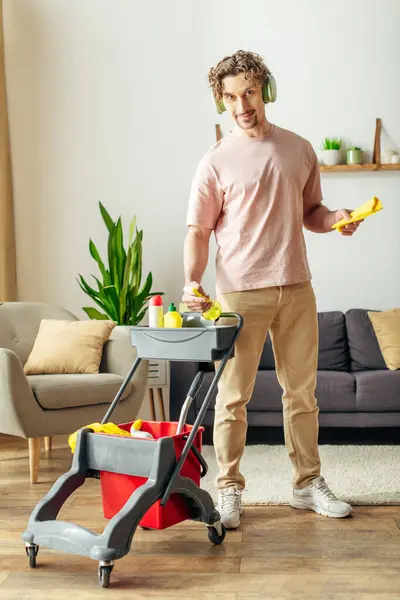 A handsome man in cozy homewear stands in a living room with a cart of cleaning supplies. — Stock Photo
