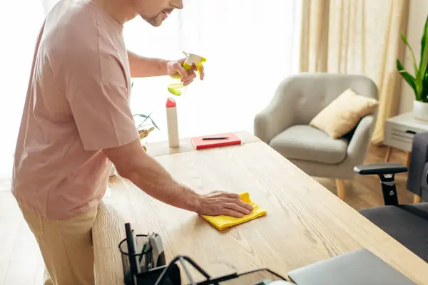 Handsome man in cozy homewear uses a yellow sponge to clean a wooden table in a sunlit room. — Stock Photo