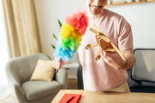 Bonito homem em casa aconchegante segurando um avião de brinquedo e espanador colorido arco-íris. — Fotografia de Stock