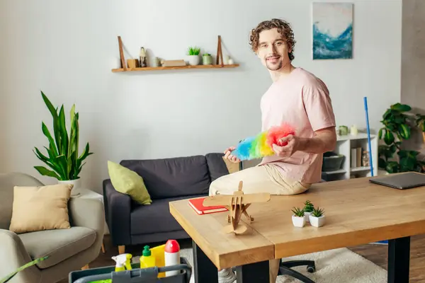 Handsome man in cozy homewear sitting on a coffee table, holding a duster. — Stock Photo