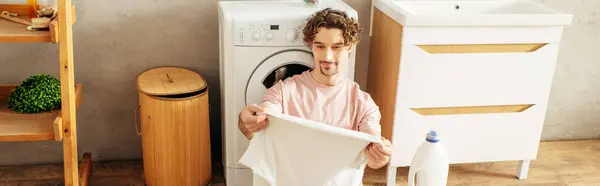 Un uomo in accogliente biancheria da casa che tiene un asciugamano di fronte a una lavatrice. — Foto stock