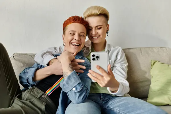 A lesbian couple with short hair sit together on a couch, smiling and taking a selfie with a phone. — Stock Photo
