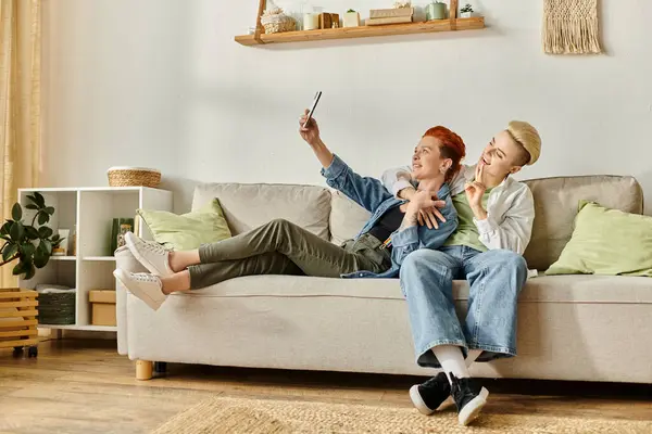 Deux femmes aux cheveux courts, un couple lesbien, assis sur un canapé prenant joyeusement un selfie ensemble. — Photo de stock