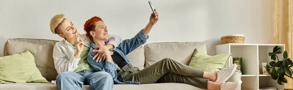 A man and woman with short hair sit on a couch, smiling and taking a selfie together in the comfort of their home. — Stock Photo