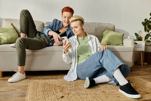 Una pareja lesbiana con el pelo corto sentado en un sofá, absorto en el teléfono inteligente - foto de stock