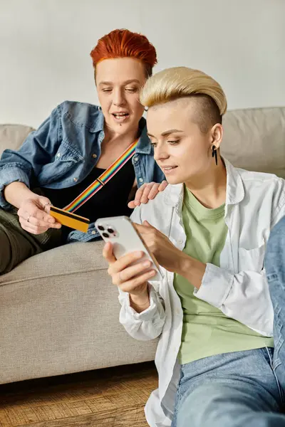 Two women, a lesbian couple, sit on a couch together looking at a cell phone screen intently. — Stock Photo