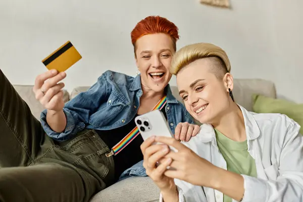 Two women, a lesbian couple with short hair, sit on a couch holding a credit card, shopping online together at home. — Stock Photo