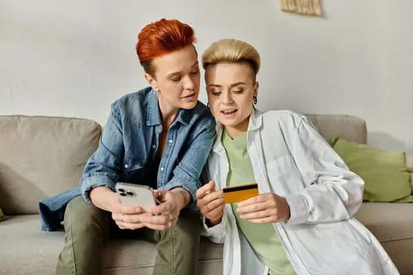 A lesbian couple with short hair sit on a couch, intently examining a credit card together. — Stock Photo