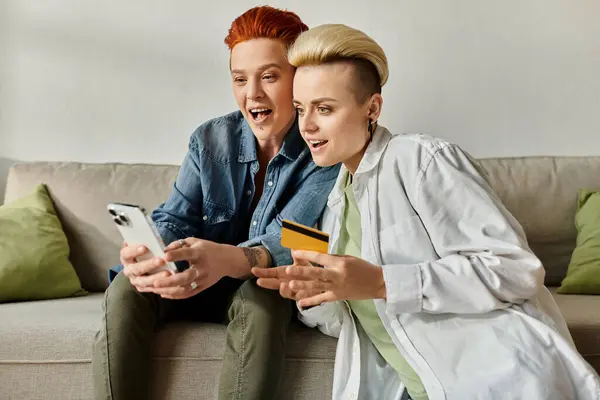 Two women with short hair sitting on a couch, contemplating a credit card in hand, in a cozy home setting. — Stock Photo