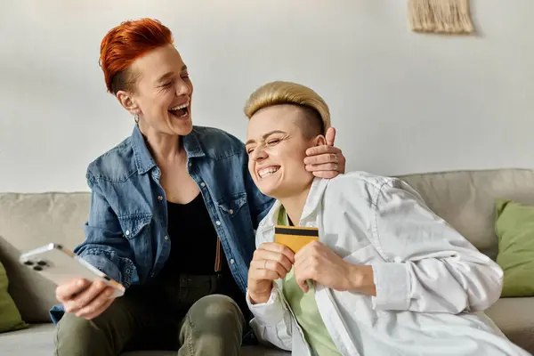 Two women with short hair sit on a couch, laughing while doing shopping online — Stock Photo